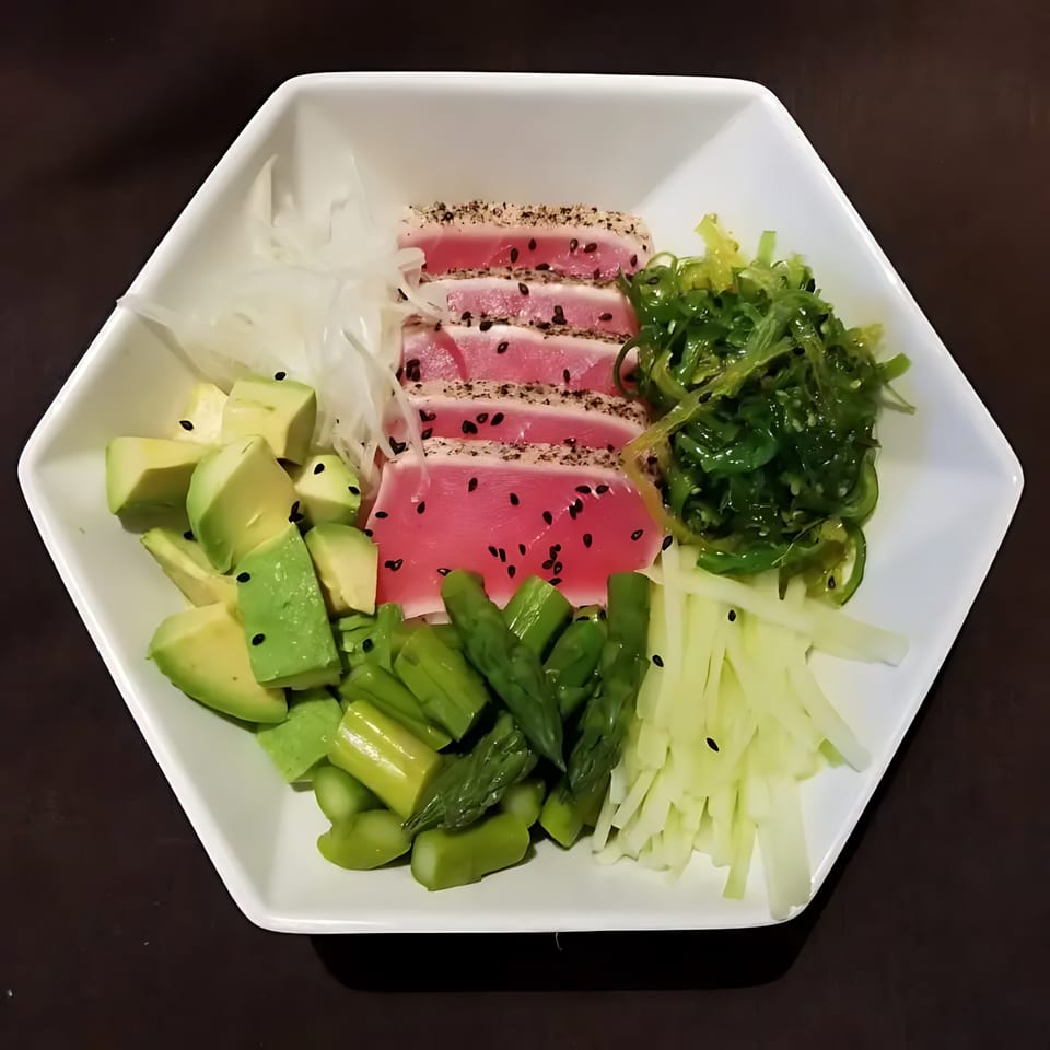 Seared tuna slices with sesame seeds, avocado, asparagus, seaweed salad, and daikon radish in a white bowl.